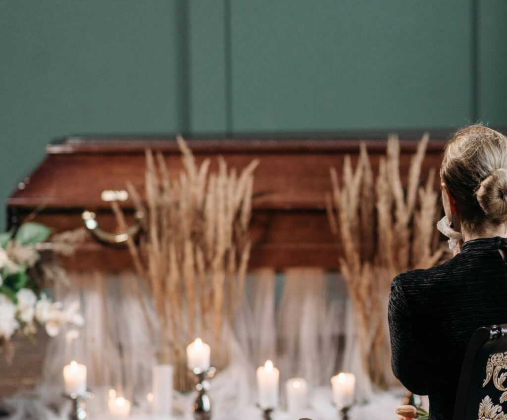 A respectful gathering of family and friends at a funeral service indoors, with floral arrangements and candles.